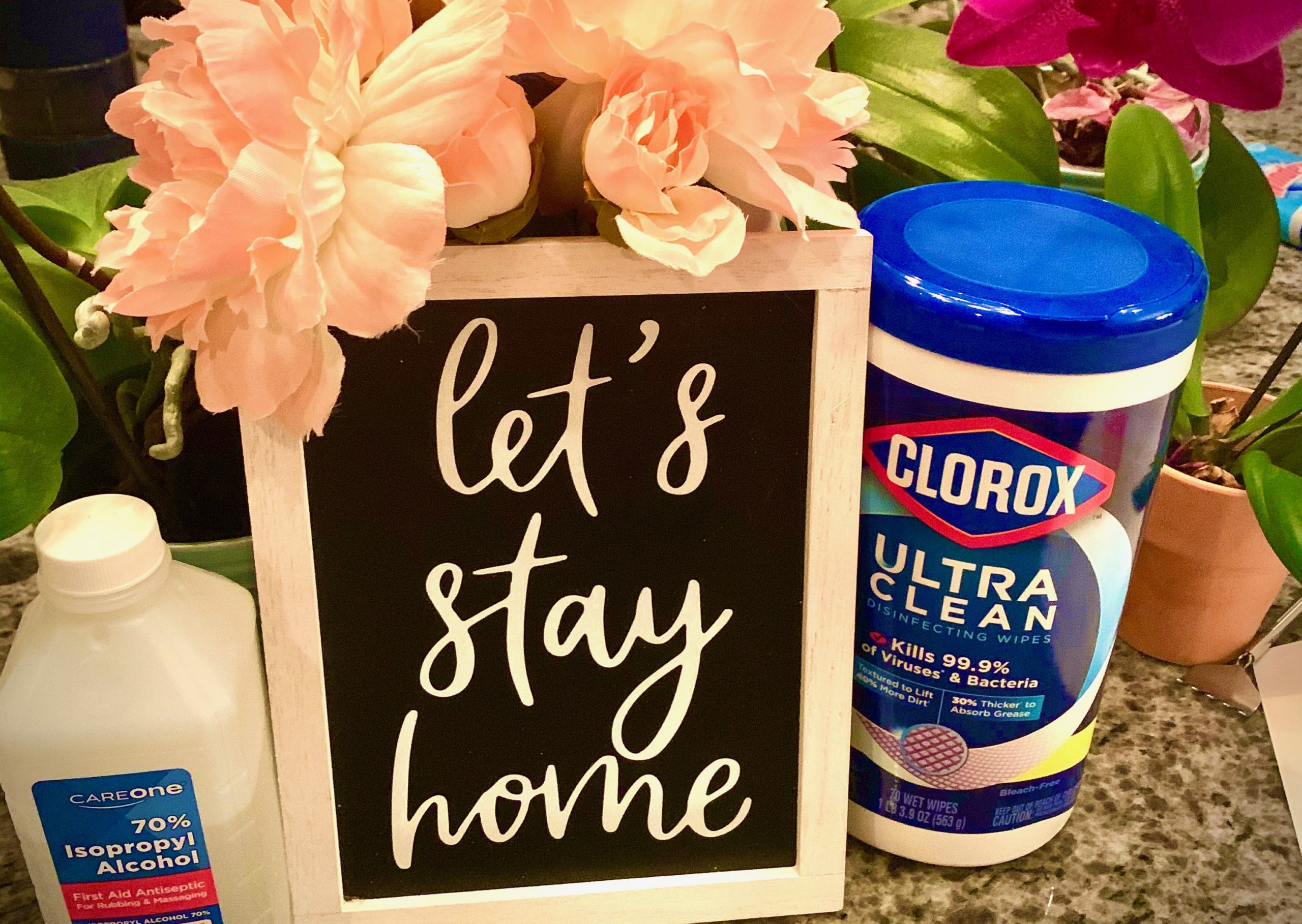 Chalkboard sign hand-lettered with "Let's Stay Home" surrounded by flowers and Clorox wipes